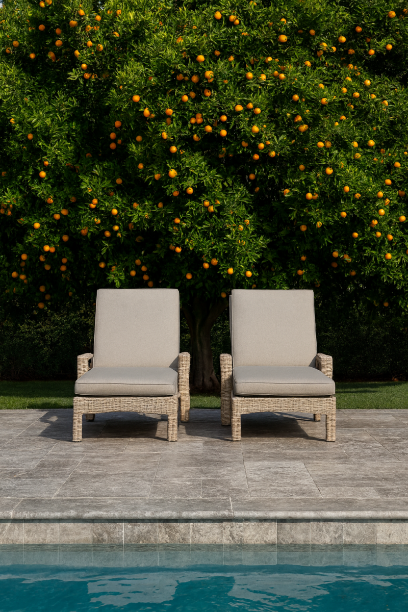 Two beige wicker lounge chairs on a marble pool deck with a clear blue pool, set against a lush green orange tree laden with fruit, conveying a serene, summery vibe.